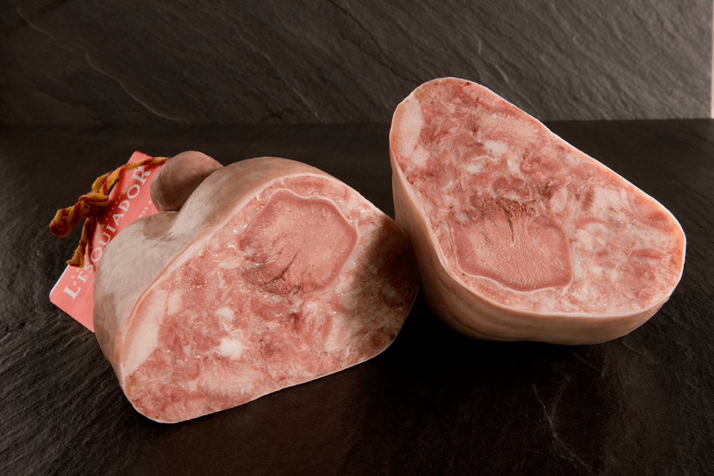 Dos piezas de cabeza de jabalí (embutido) cortadas por la mitad, mostrando una mezcla de carne rosada y partes blancas, con una etiqueta roja de la marca "L'Esquador" en el fondo.